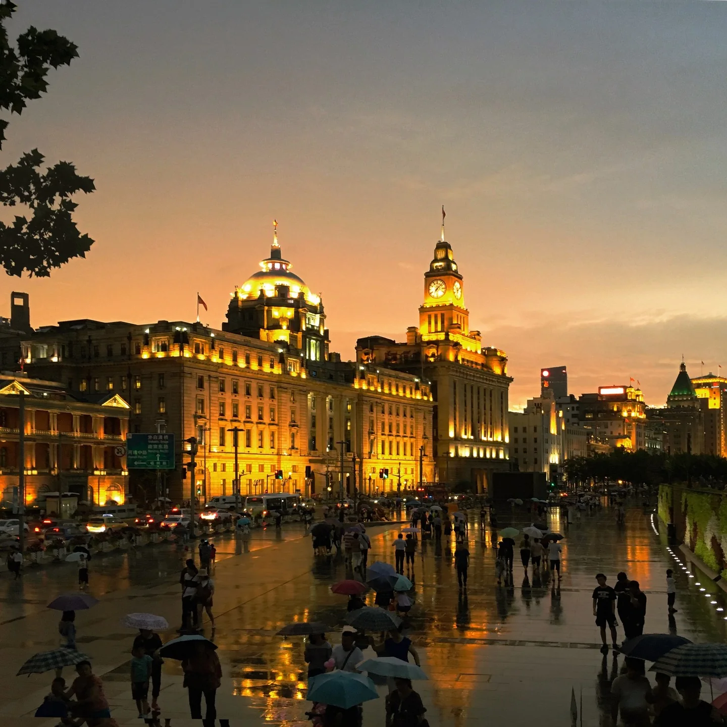 View of historic buildings on the Bund. The area is freshly wet from rain. People have umbrellas and the wet sidewalks reflect the warm-colored lights from the buildings.