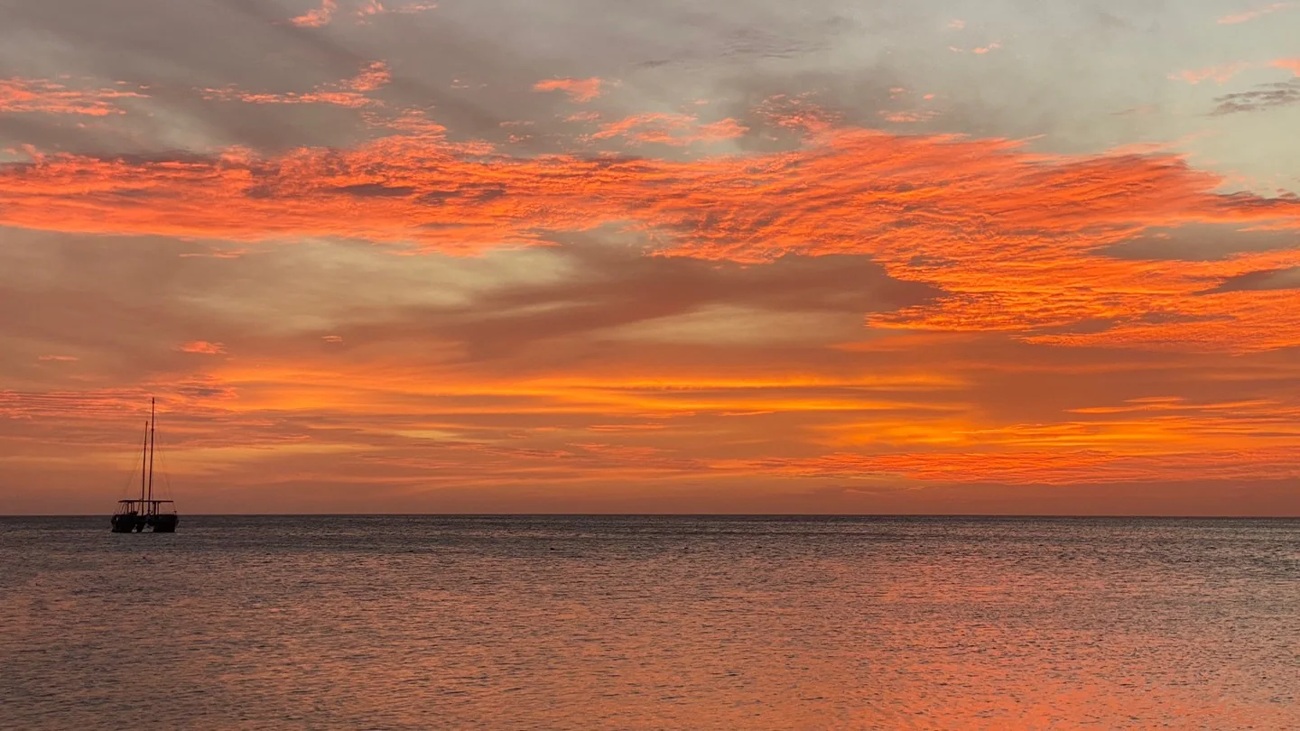 Fiery sunset over calm ocean, a small boat on the water is silhouetted against the sky.