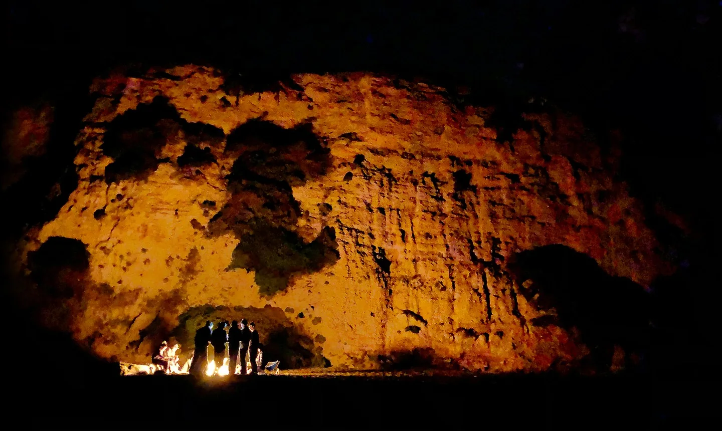 A group around a campfire at night with shadows cast on the cliff face behind them.